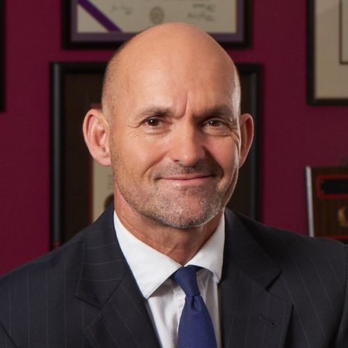 Bald man in a suit and tie smiling, with framed certificates on a maroon wall background.