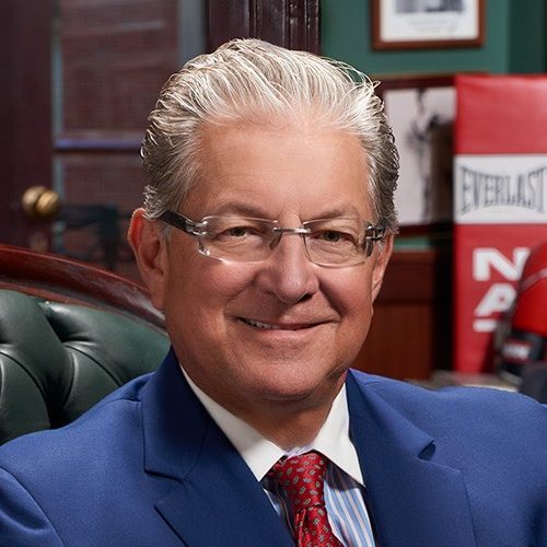 Smiling man with glasses in a blue suit and red tie, seated in an office with boxing memorabilia in the background.