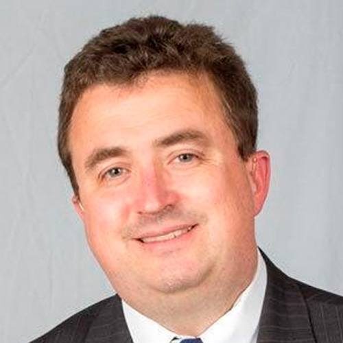 Smiling man in a suit and tie with short, curly hair against a plain background.
