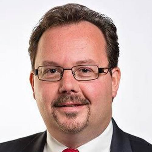 Man with glasses, short dark hair, mustache, and goatee, wearing a suit and red tie, smiling slightly.