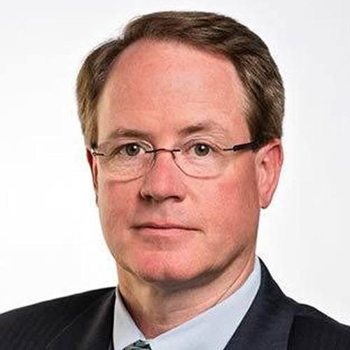 Man with glasses, brown hair, wearing a suit and tie, neutral expression, white background.