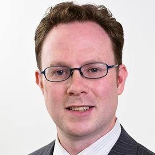 Man with short hair, glasses, and a suit, smiling against a plain background.