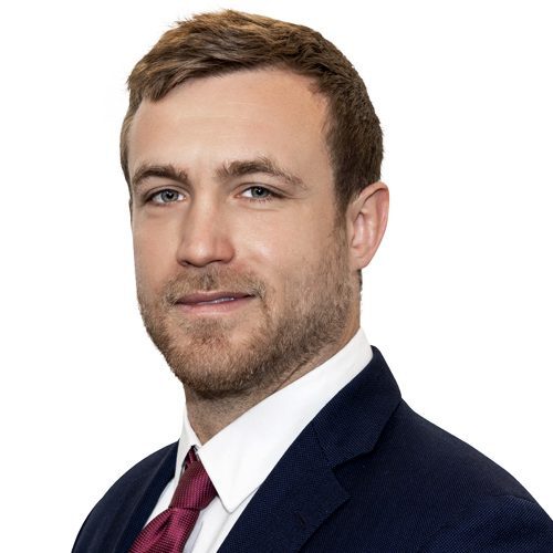 Man with short hair, beard, wearing a navy suit, white shirt, and red tie, looking at the camera against a white background.