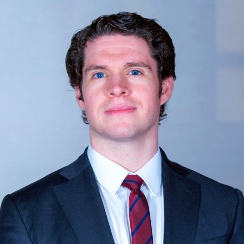 Man in a suit with a red striped tie, dark curly hair, and blue eyes, against a light gray background.