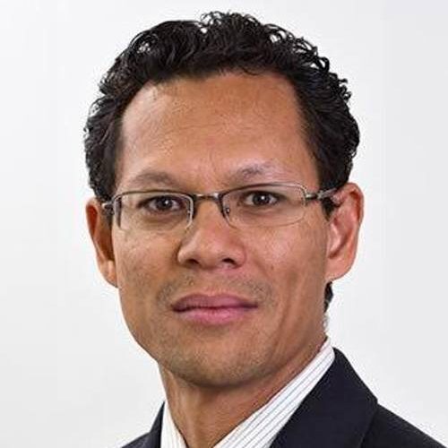 Man with glasses, dark curly hair, wearing a suit and tie, against a plain white background.