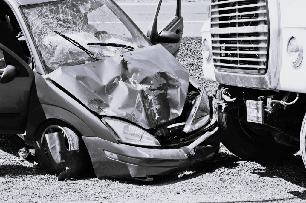 Crashed car with a smashed front end next to a large truck on a gravel road.